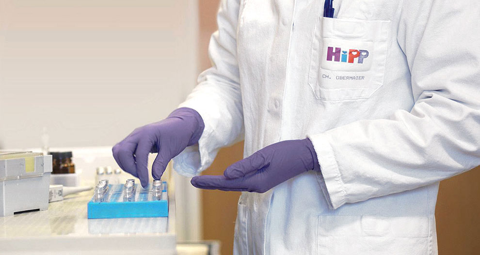 Лице в бяла лабораторна престилка с логото "HiPP" и името "CH. OBERMAIER" е с лилави ръкавици и борави със синя табла с множество малки контейнери.