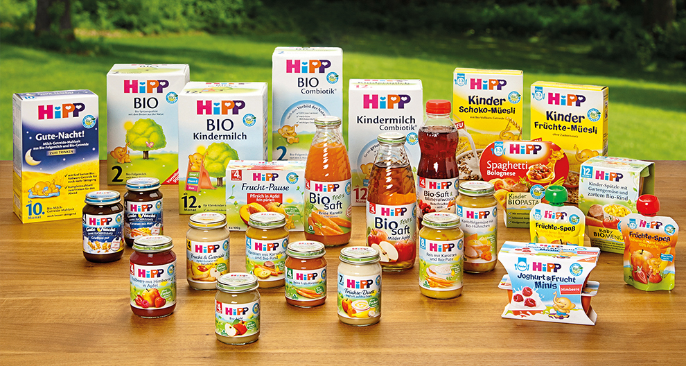 Различни продукти за бебешка храна Hipp, изложени на маса на зелен фон на открито.