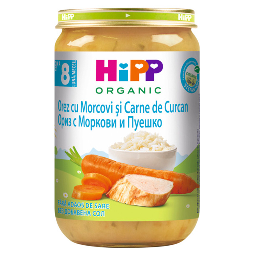 Бурканче с органична бебешка храна Hipp с етикет на два езика, на който са изобразени морков и парче месо, на фона на тревисто поле и купичка с ориз.