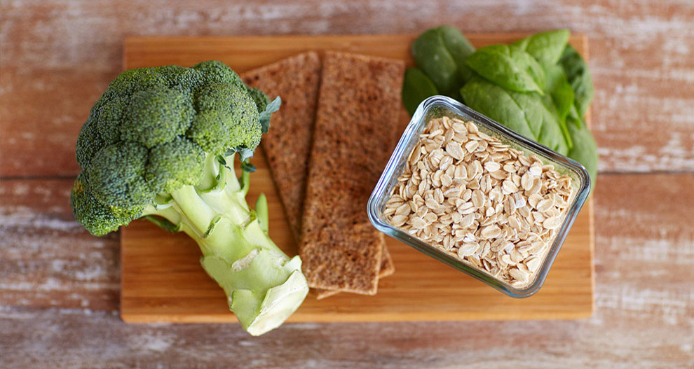 A wooden cutting board holds a stalk of broccoli, a bunch of spinach, a rectangular glass container of rolled oats, and two rectangular brown crackers.
