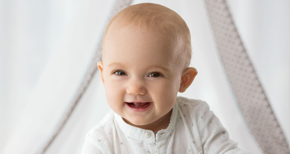 A baby with a white outfit and a dotted pattern is smiling, with a white curtain in the background.
