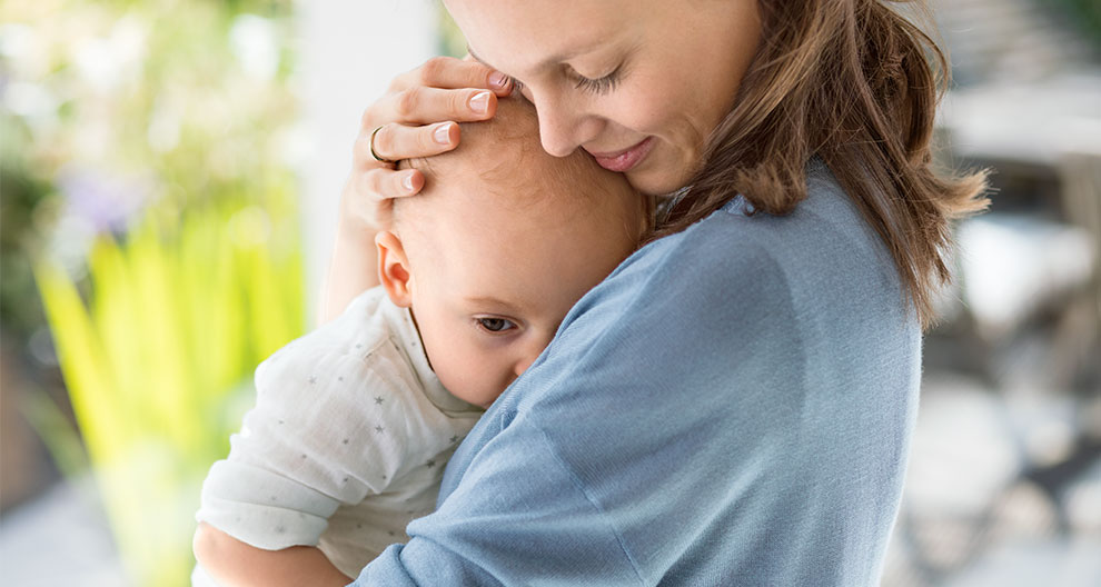 A woman in a blue shirt holds a baby in a white outfit, both facing the camera.