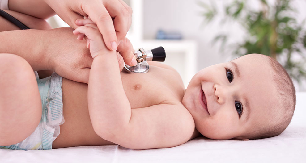A baby lies on a white surface, wearing a light blue diaper, while a stethoscope is placed on the baby's chest by a hand.
