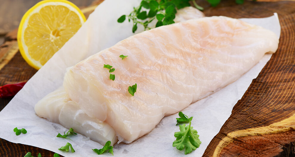 A fresh fish fillet rests on a wooden surface, accompanied by a slice of lemon and a sprig of parsley.
