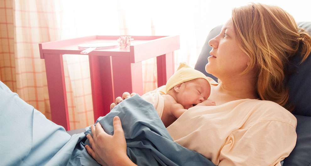 A woman reclines on a bed, holding a newborn baby wrapped in a blue blanket.