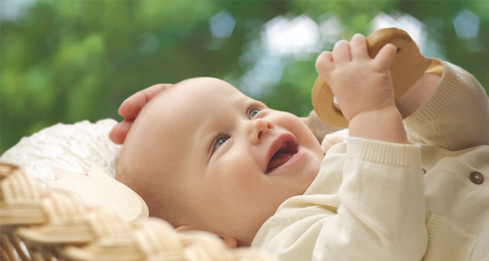 A baby lies in a basket, holding a wooden toy, with a blurred green background.