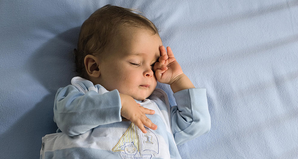 A baby lies on a blue surface, wearing a light blue outfit with a sailboat design, and appears to be in a state of rest or sleep.