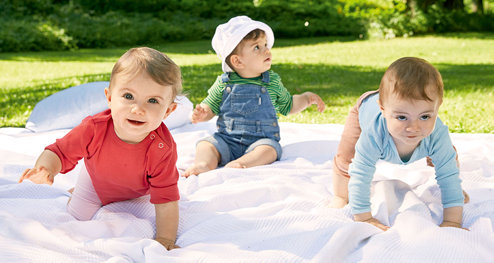 Three toddlers are playing on a white blanket in a grassy area.