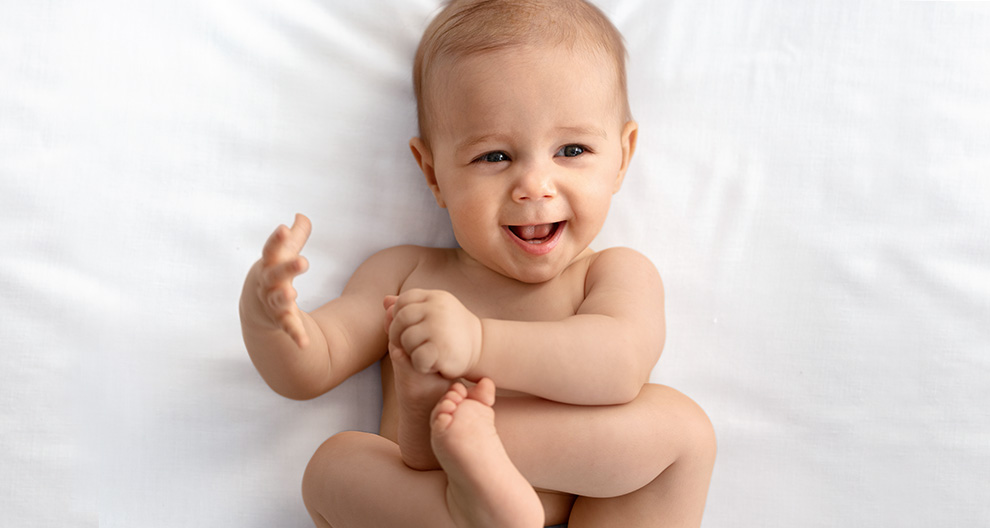 A baby lies on a white surface, reaching out with one hand.