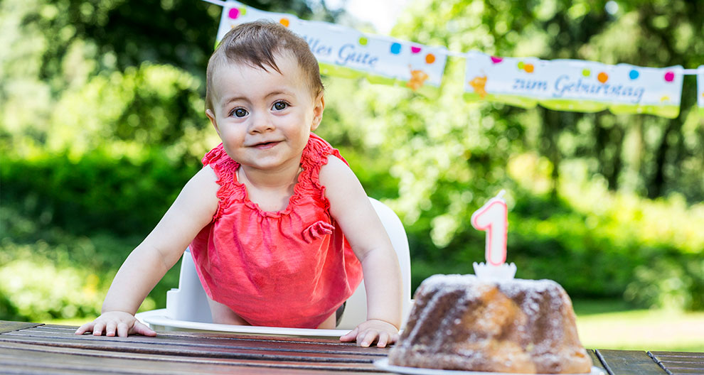 A baby in a red dress sits at a table with a cake featuring a number one candle.