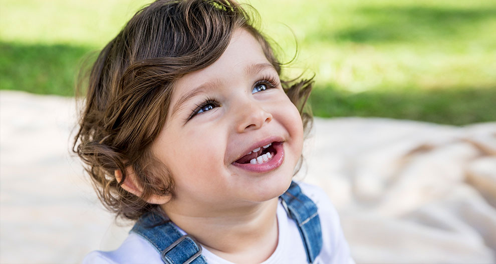 A child with curly hair, wearing a white shirt and blue suspenders, looking up with a smile.