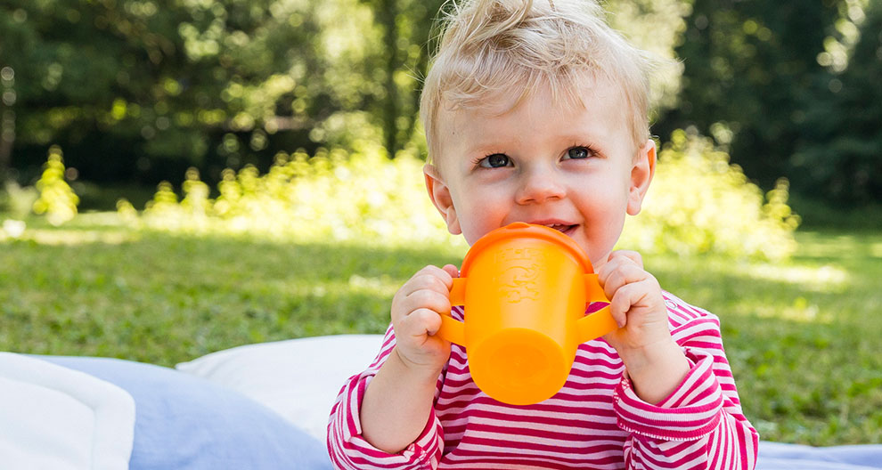 A toddler sits outdoors on a blanket, holding an orange cup.