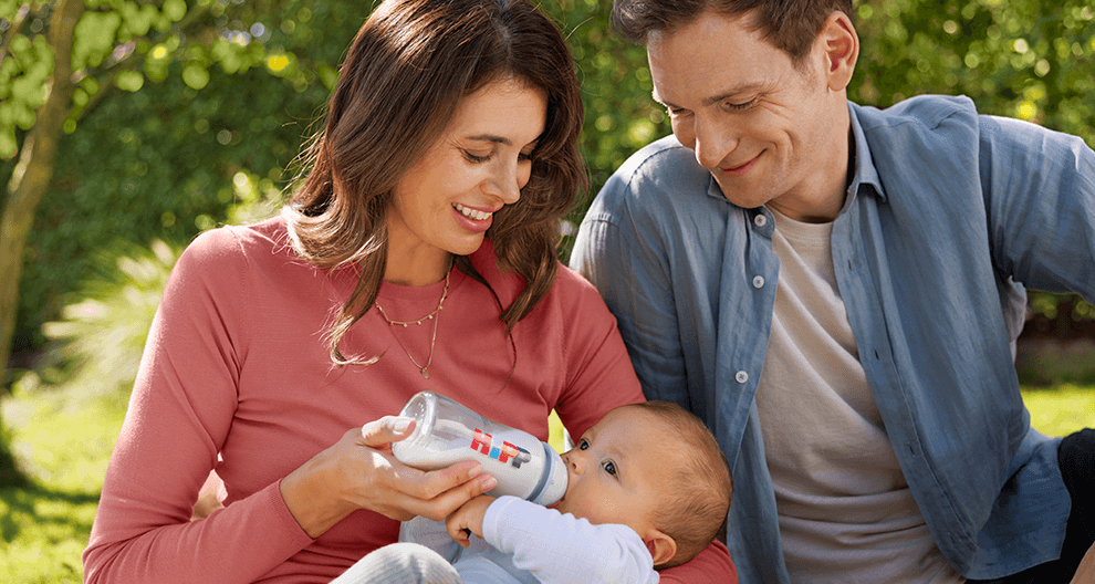 A man and a woman sit outdoors, feeding a baby with a bottle.