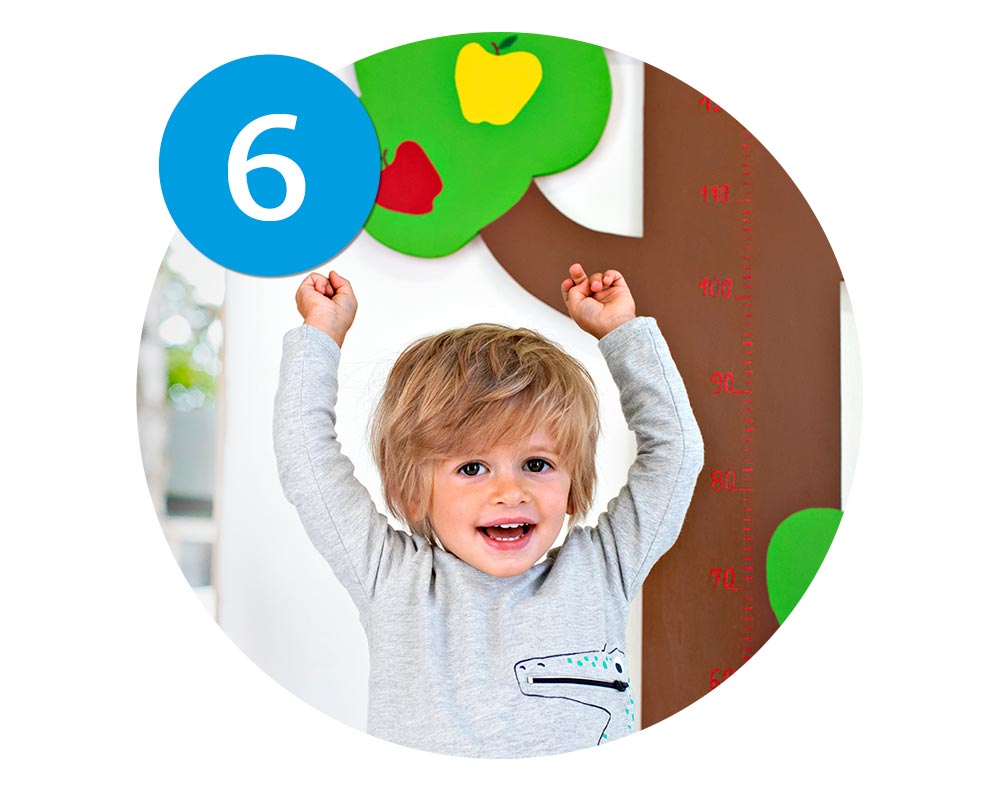 A child stands in front of a height measurement chart, raising their arms in excitement.